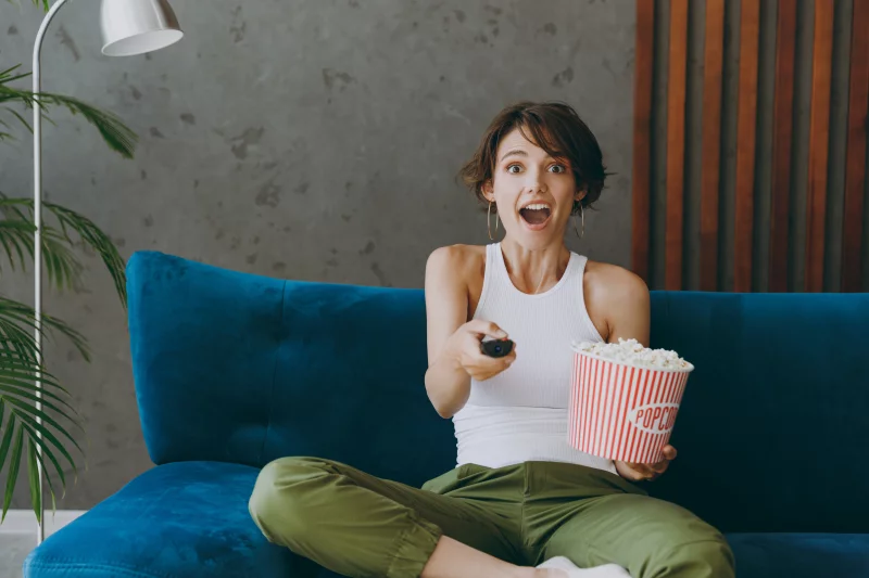 Young shocked woman in white tank shirt hold TV remote controller pop corn watch film switch channels sit on blue sofa stay at home hotel flat rest relax spend free spare time in living room indoors